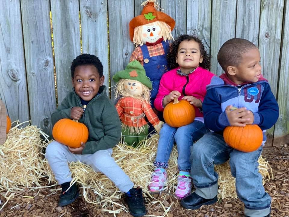 Happy little young preschool kids holding a pumpkin while sitting on wood chips on a scarecrow background at a Preschool & Daycare Serving Hampton Roads, VA