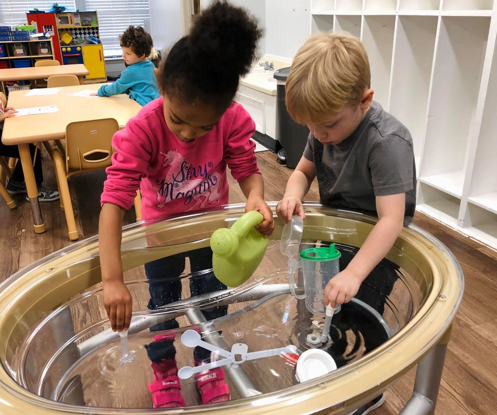 A little preschool girl and a boy enjoying their water activity on a clear water table with accessories at a Preschool & Daycare Serving Hampton Roads, VA