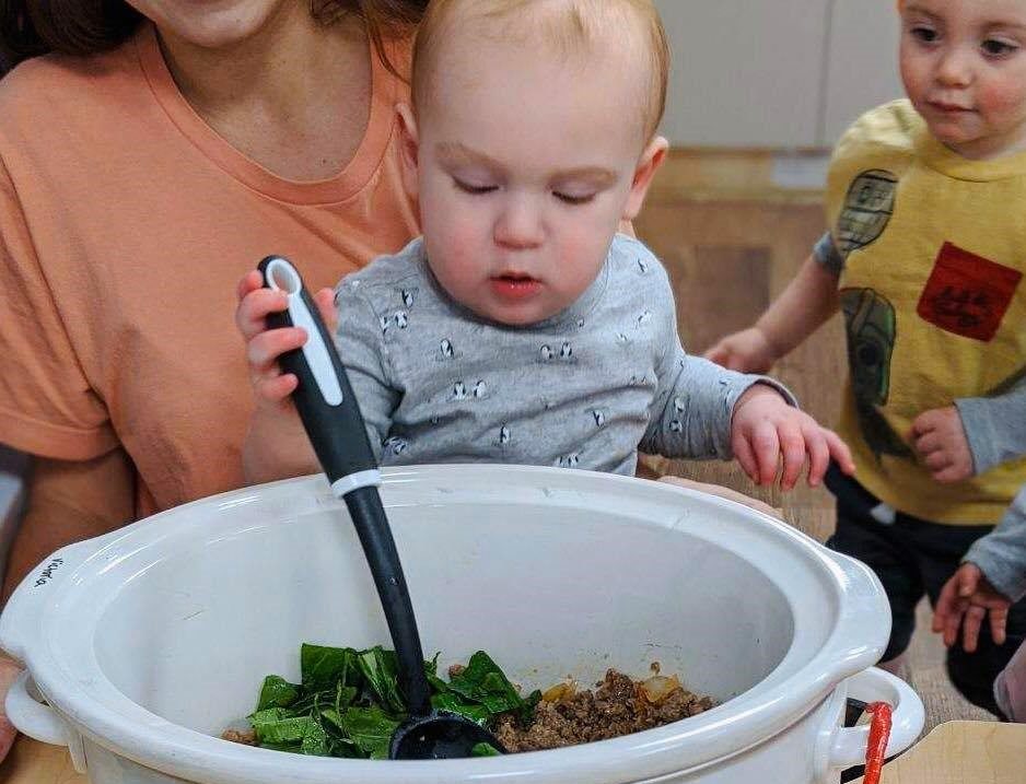 A toddler being watched by another one and a teacher mixing some leafy vegetables and cooked ground pork in a bowl using a spatula at a Preschool & Daycare Serving Hampton Roads, VA